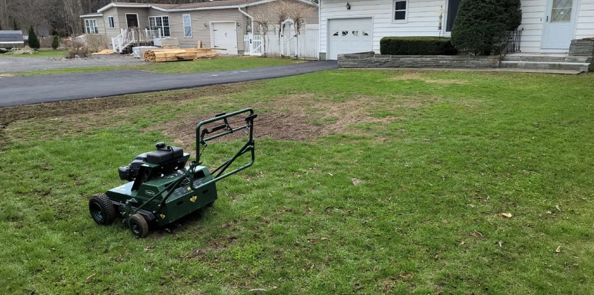A front lawn, aerated and overseeded in spring. The aerator is in the before photo. Before: A thin front lawn with bare patches and a walk-behind aerator parked on it, in front of a white house.