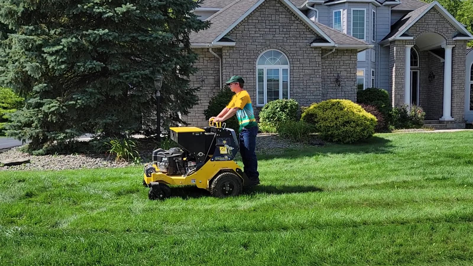 A technician in the Happy Habitat uniform riding a yellow stand-on aerator, with a seed spreader on board, across a green front lawn.