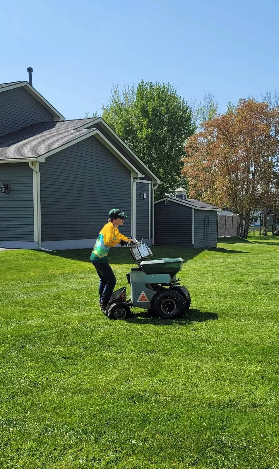 A Happy Habitat technician running a stand-on spreader across a lawn beside a gray home.